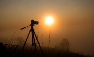 Silhouette of photo camera with tripod on sunrise in the fog, outdoors